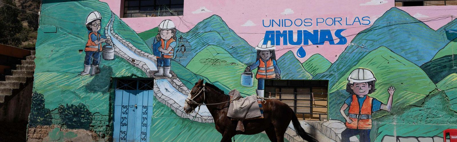 a saddled brown horse stands outside of a building with a painted mural of green mountains, a pink sky, and people in construction garb standing along a canal under the words "Unidos por las amunas".